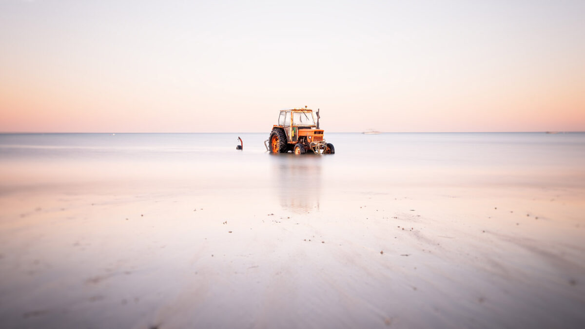 Tracteur orange stationné sur la plage d'Agon-Coutainville à marée montante, avec un reflet doux sur l'eau lisse et un ciel clair de fin de journée en Normandie.