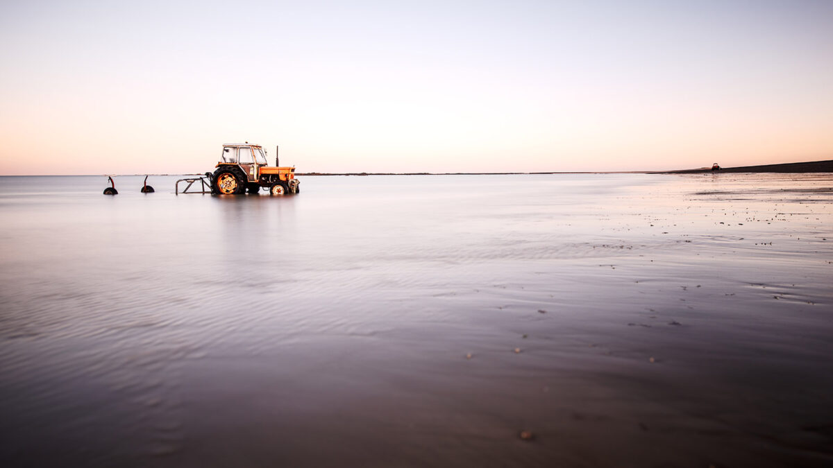 Tracteur orange stationné sur la plage d'Agon-Coutainville à marée montante, avec un reflet doux sur l'eau lisse et un ciel clair de fin de journée en Normandie.